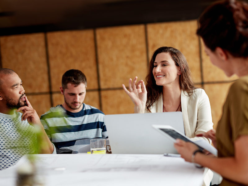 Group of professionals in a meeting room. A woman smiles and gestures while speaking, others listen attentively. Laptops and papers are on the table.