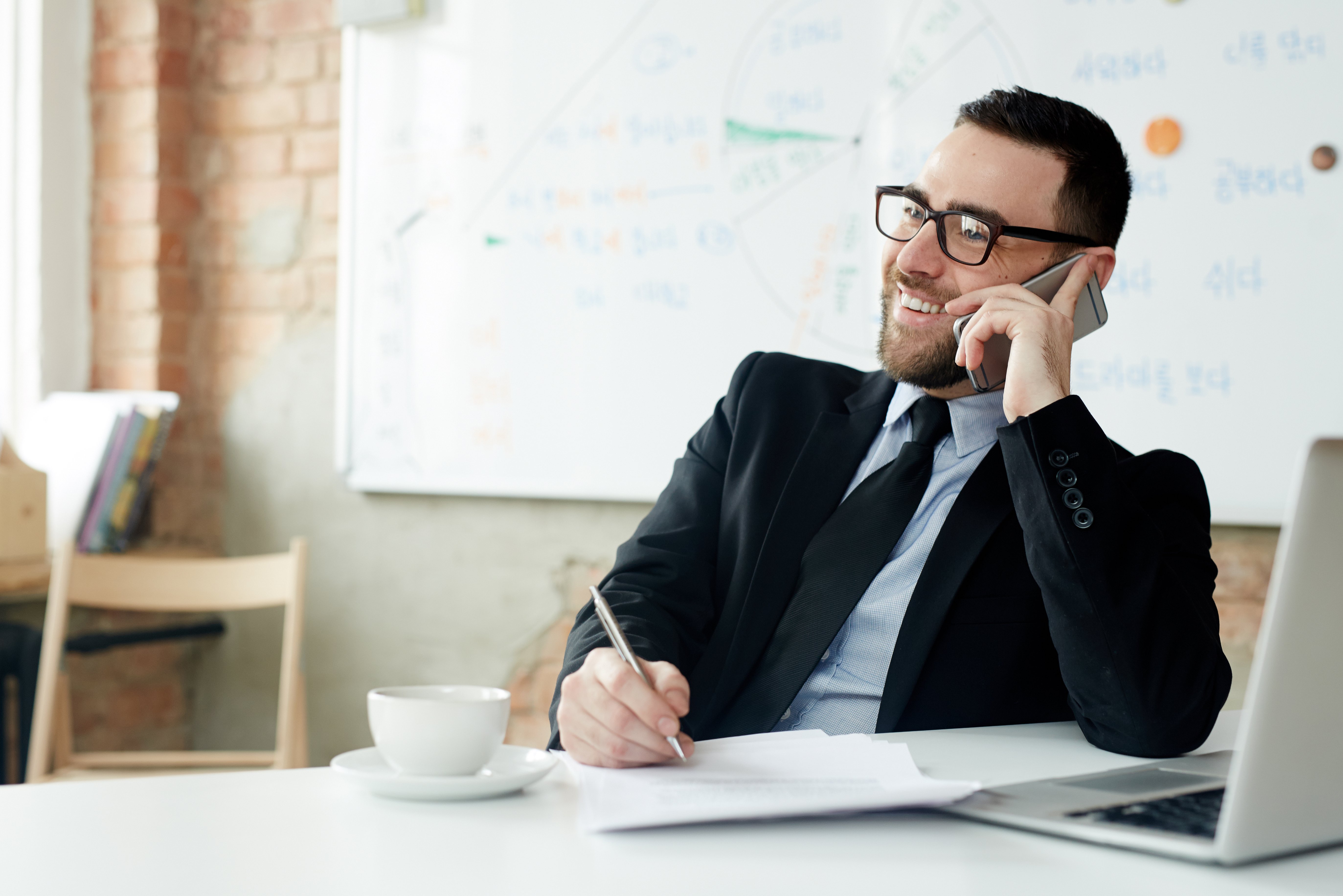 A man in a suit smiles while talking on a phone, jotting notes at a desk with papers and a laptop. A coffee cup and whiteboard are in the background.