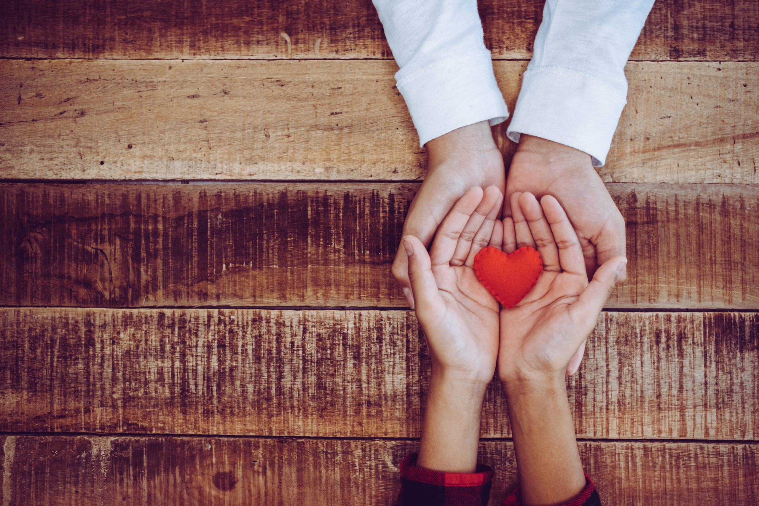 Two pairs of hands holding a red heart together, showing vulnerability.