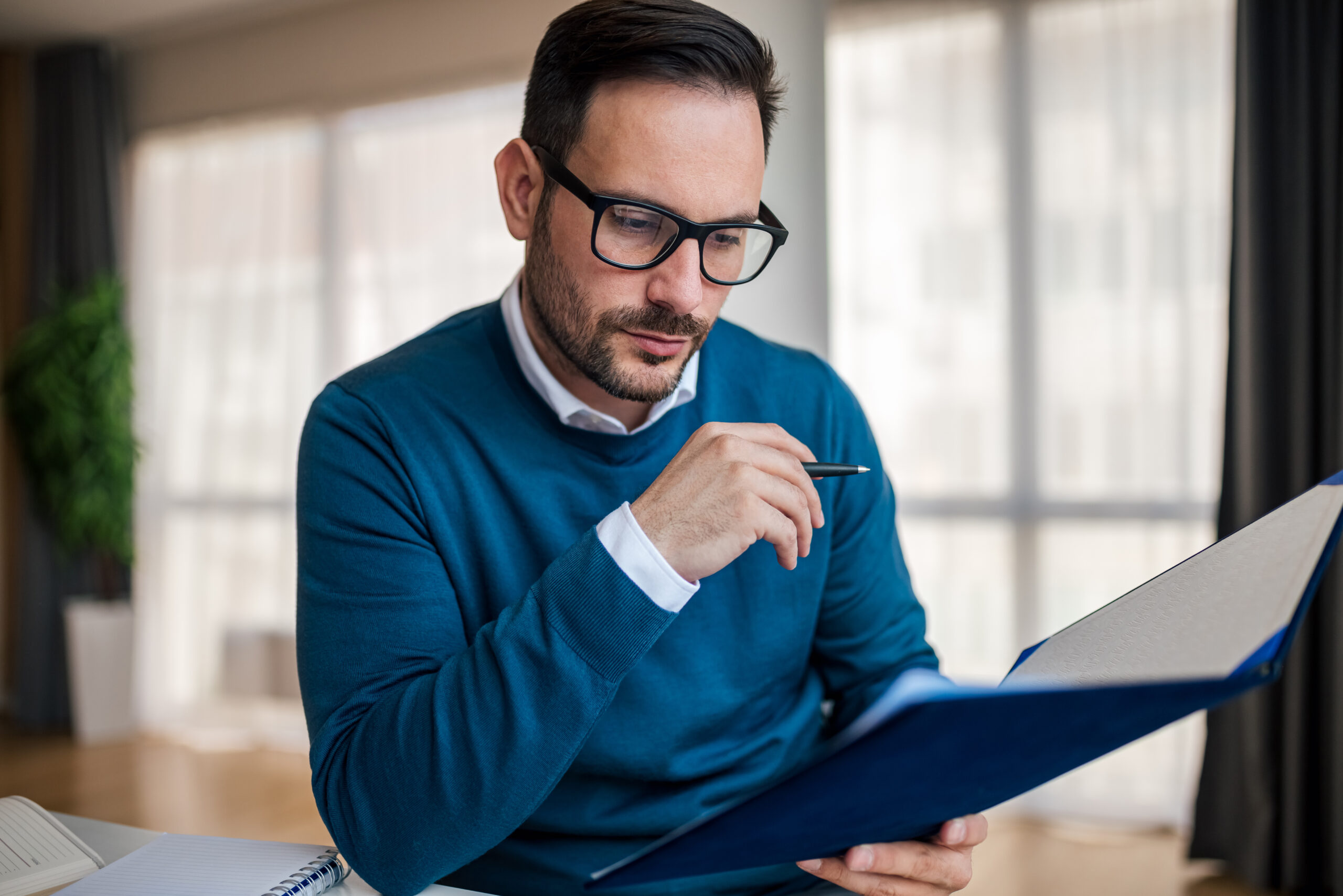 Businessman wearing glasses holds a pen and folder while reviewing documents contained therein. [What’s the Antidote to Frustration? Persistence. ]