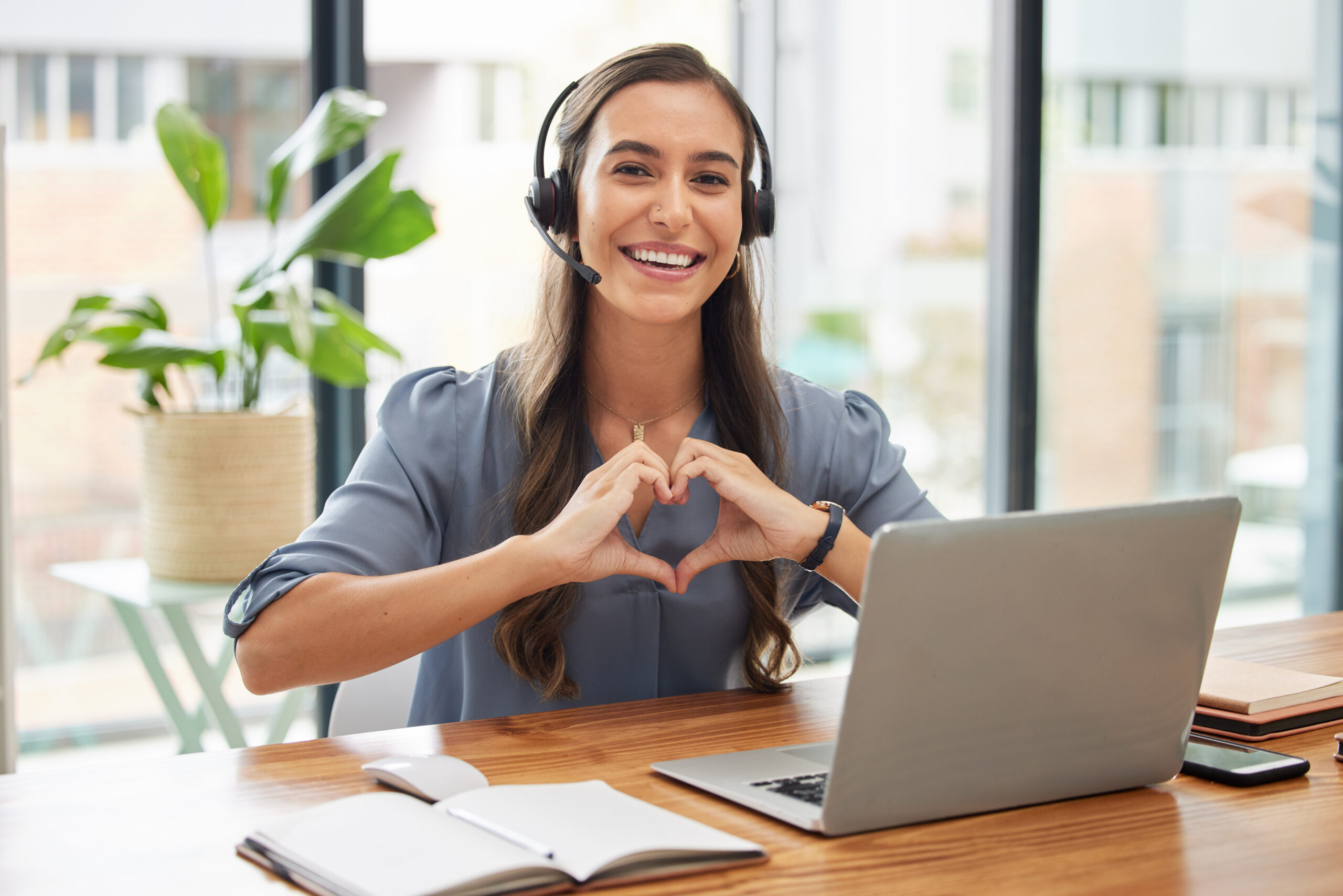 A woman with a headset sits in front of a laptop, making the shape of a heart with her hands [10 Ways to Wow Your Major Donors and Win Them for Life]