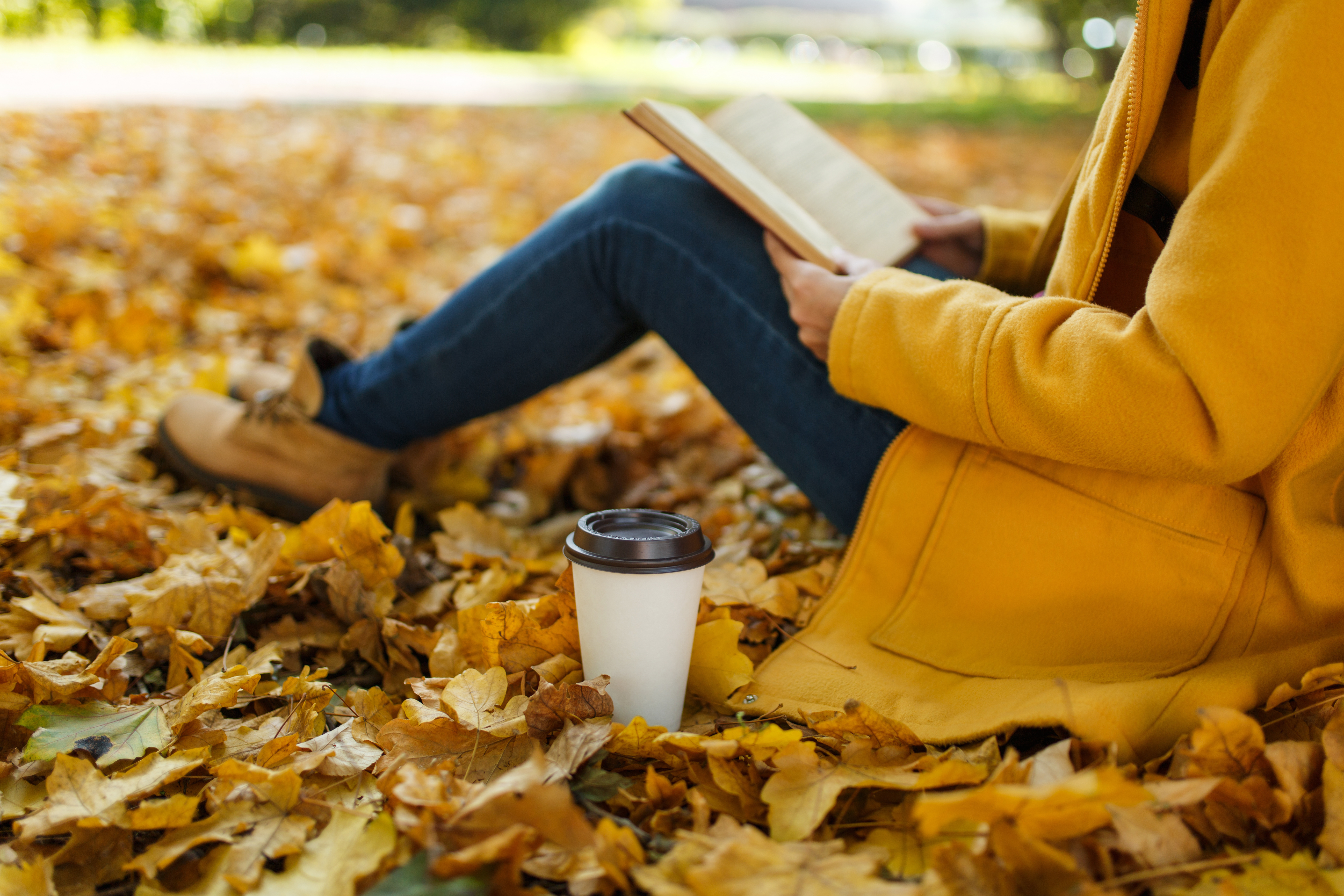 Woman in yellow coat, jeans and boots sitting under maple tree with a journal and cup of coffee in city park in Fall. [Fall is the Perfect Time to Replenish Your Why]